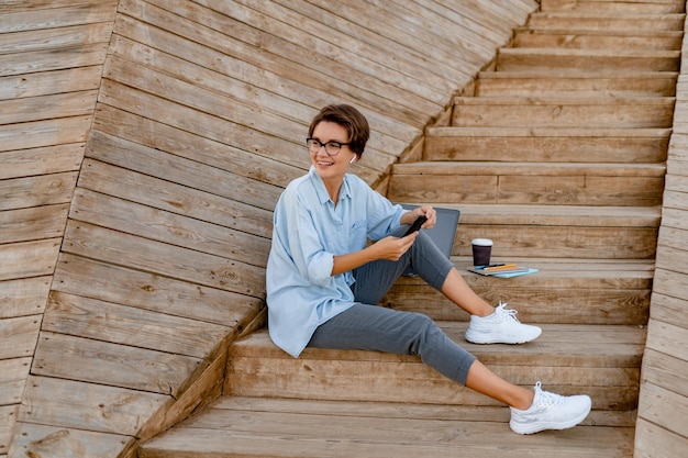 Free photo young pretty woman sitting with laptop in modern park street using smartphone