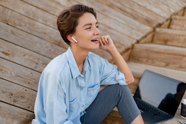 Young pretty woman sitting with laptop in modern park street using smartphone