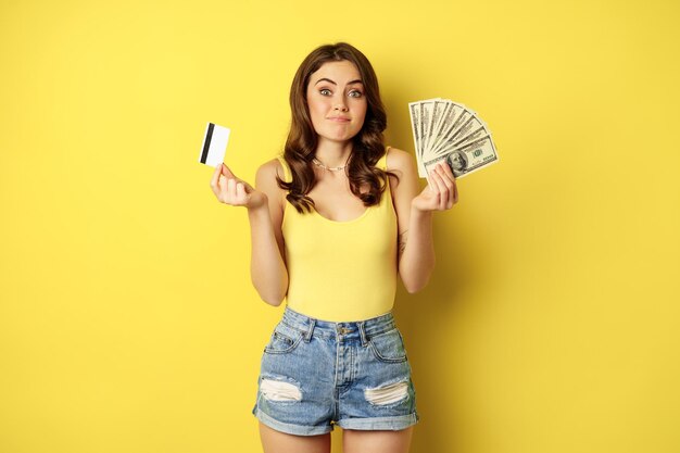 Молодая красивая женщина в летнем наряде держит в руках кредитную карту и деньги наличными, стоя против желт...