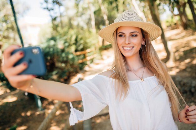 公園でスタイリッシュな夏帽子でselfieを取る若いきれいな女性。