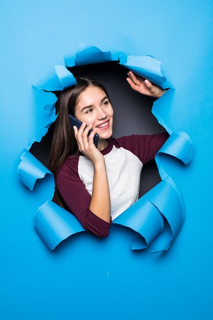 Молодой милый телефон беседы женщины пока смотрящ через голубое отверстие в бумажной стене.