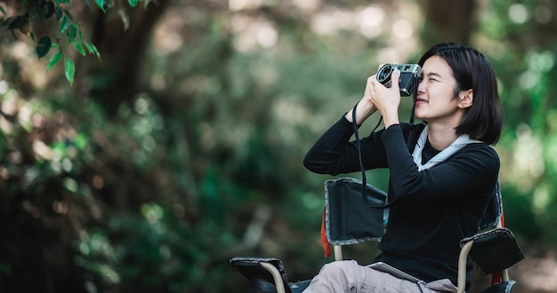 젊은 예쁜 여성은 행복 복사 공간이 있는 숲에서 캠핑하는 동안 아름다운 자연을 사진으로 찍는 디지털 카메라를 사용합니다