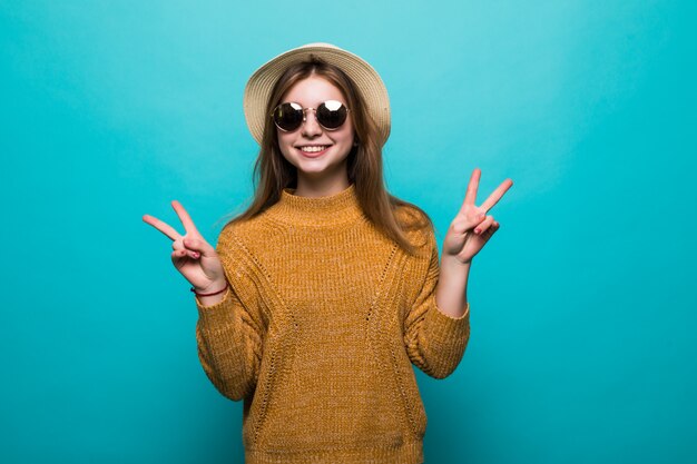 Young pretty woman wearing in hat and sunglasses pointed peace gesture on blue wall