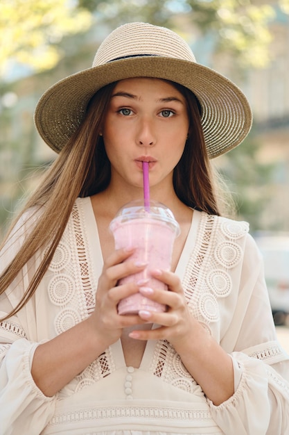 Young pretty woman in white dress and hat drinking smoothie dreamily looking in camera while standing on city street alone