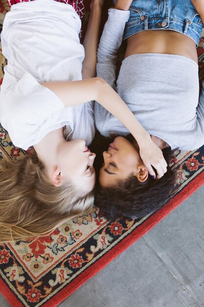 Free photo young pretty woman with blond hair and nice african american woman with dark curly hair dreamily lying on vintage carpet while spending time together at home
