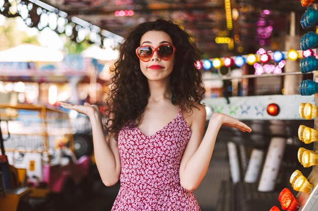 Young pretty woman with dark curly hair in dress standing and showing hands to the side while thoughtfully looking in camera in amusement park