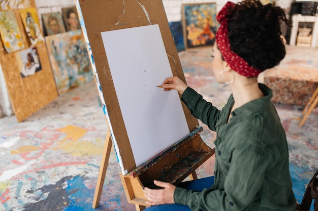 Free photo young pretty woman with dark curly hair from back sitting on chair dreamily drawing picture on canvas while spending time in cozy art workshop