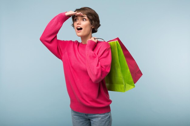 Молодая красивая женщина с темными короткими волосами в розовом свитере и джинсах, держащая на плече красочные сумки для покупок, изумленно глядя в сторону на синем фоне