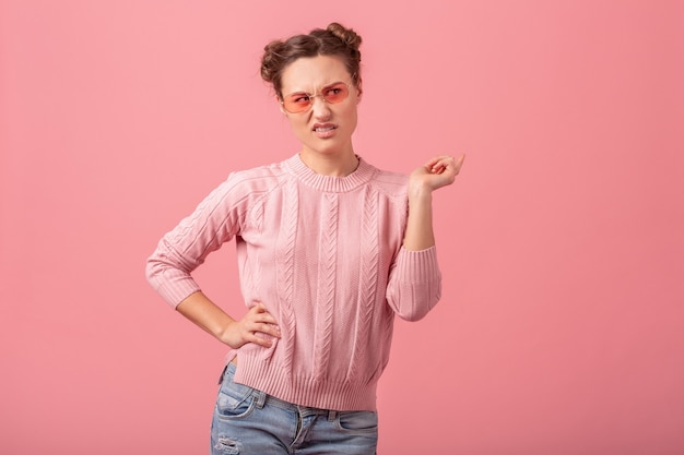 Young pretty woman with funny face expression, poiting finger, feeling disgust, in pink sweater and sunglasses isolated on pink studio background