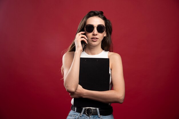 Young pretty woman with glasses speaking on phone and holding notebooks 