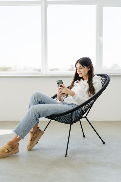 Молодая красивая женщина с мобильным телефоном сидит в кресле папасан возле окна дома