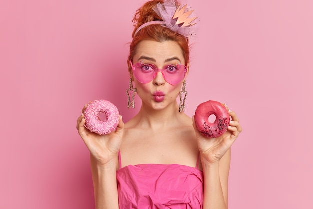 Free photo young pretty woman with red hair has sweet tooth keeps lips folded holds two tasty doughnuts