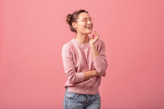 Young pretty woman with sincere candid smile in pink sweater and sunglasses isolated on pink studio background