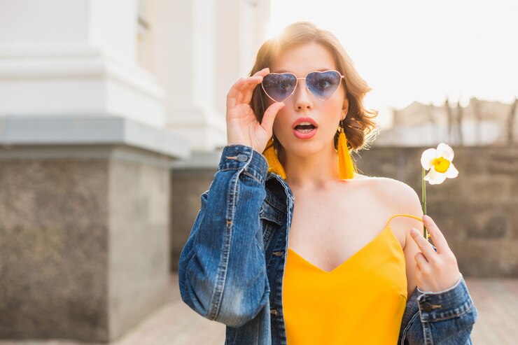 Free Photo young pretty woman with surprised face expresssion, emotional, shocked emotion, wearing stylish apparel, denim jacket, yellow top, holding flower, sunny summer, trendy funny blue sunglasses