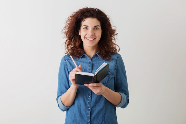 Young pretty woman writing in notebook with pencil, smiling, curly hair, positive, happy, isolated, denim blue shirt, hipster style, student learning, looking in camera, making notes, education