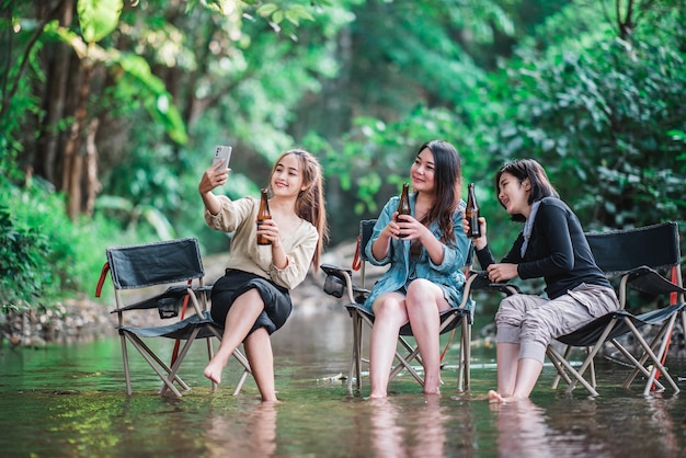 맥주병을 들고 개울의 캠핑 의자에 앉아 스마트폰 셀카를 행복으로 사용하는 젊고 예쁜 여성들