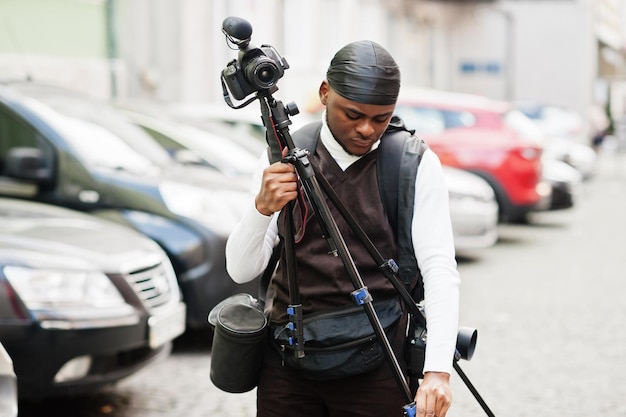 Foto gratuita giovane videografo afroamericano professionista che tiene una fotocamera professionale con attrezzatura pro treppiede cameraman afro che indossa duraq nero che realizza un video
