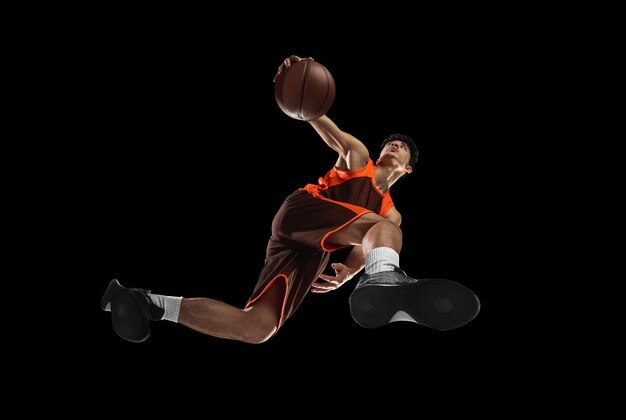 Молодой профессиональный баскетболист в действии, движение, изолированное на черной стене, взгляд снизу. Понятие спорта, движения, энергии и динамичного, здорового образа жизни. Обучение, практика.
