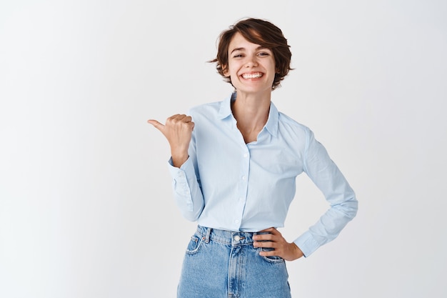 Young professional ceo woman in blue collar shirt, smiling happy and pointing left at, showing promo, standing on white wall