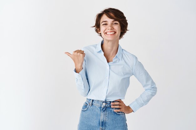 Молодой профессиональный генеральный директор женщина в синей рубашке с воротником, счастливая улыбающаяся и указывая влево, показывая промо, стоя на белой стене