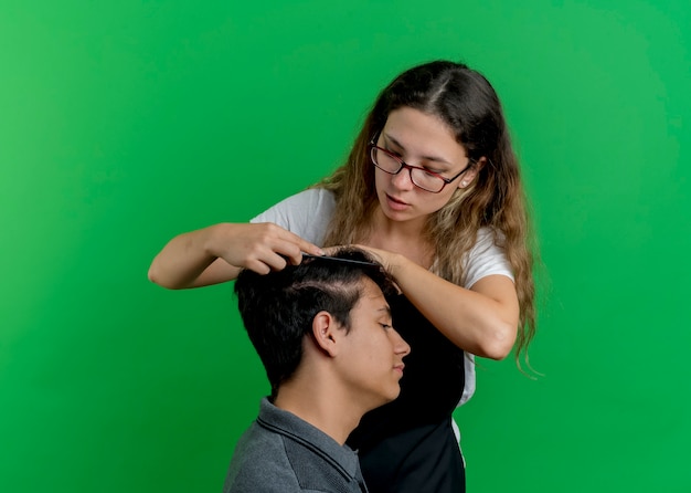 Young professional hairdresser woman in apron combing hair of man client standing over green wall