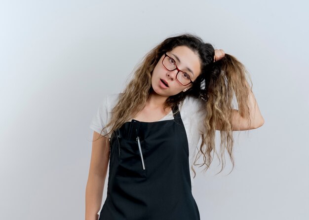 Young professional hairdresser woman in apron confused touching her hair 