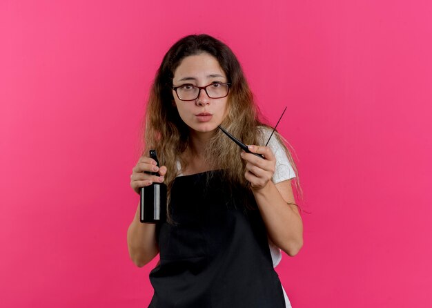 Young professional hairdresser woman in apron holding comb and spray lookign at camera with serious face 