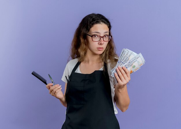 Молодой профессиональный парикмахер женщина в фартуке держит в замешательстве расческу для волос, ножницы и деньги