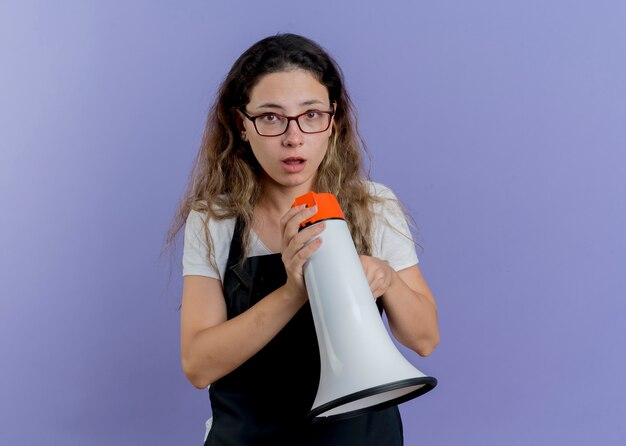 Young professional hairdresser woman in apron holding megaphone looking at front confused standing over blue wall