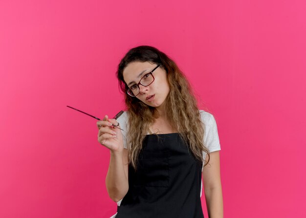 Молодой профессиональный парикмахер женщина в фартуке, держа ножницы и расческу с серьезным лицом
