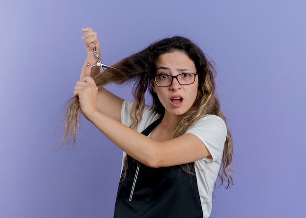 Молодой профессиональный парикмахер женщина в фартуке, держащая ножницы, стригущая волосы, выглядит смущенной