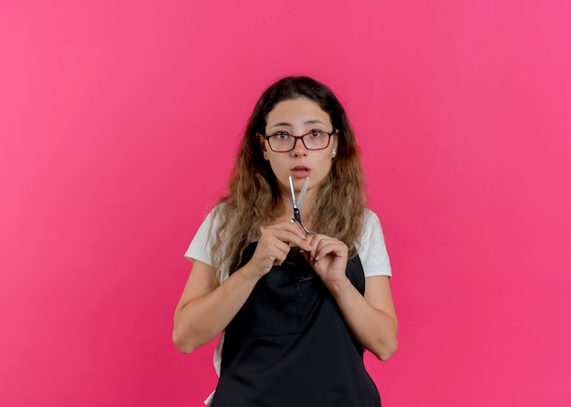 Free photo young professional hairdresser woman in apron holding scissors looking at front being confused standing over pink wall