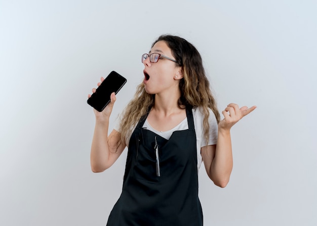 Young professional hairdresser woman in apron holding smartphone using it as microphone singing having fun standing over white wall