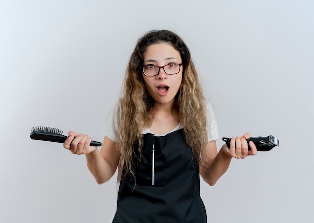 Young professional hairdresser woman in apron holding trimmer and hair brush looking at front surprised and confused standing over white wall