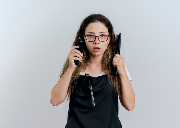 Young professional hairdresser woman in apron holding trimmer and hair brush looking at front with serious face standing over white wall