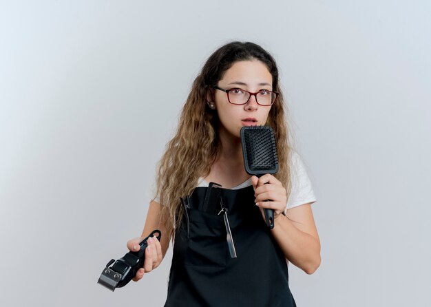 Young professional hairdresser woman in apron holding trimmer and hair brush looking at front with serious face standing over white wall