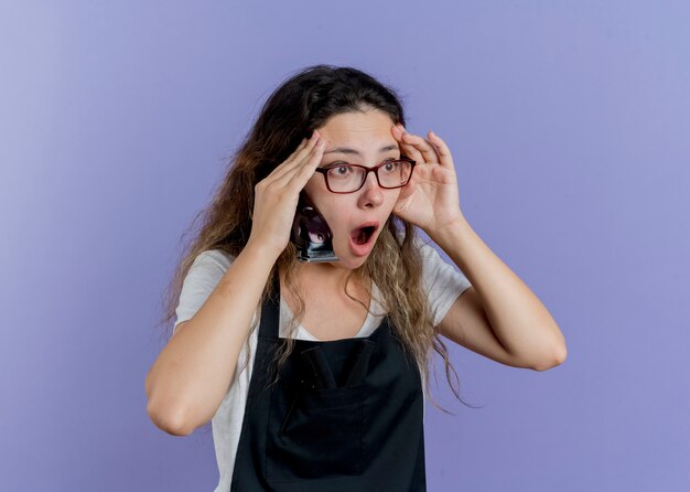 Young professional hairdresser woman in apron holding trimmer looking aside being in panic 