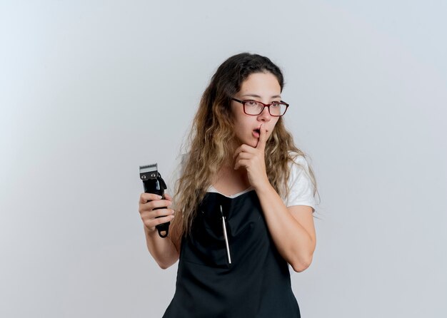 Young professional hairdresser woman in apron holding trimmer looking aside puzzled standing over white wall