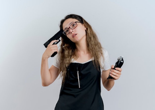 Young professional hairdresser woman in apron holding trimmer and pistol near her temple looking aside tired and bored standing over white wall