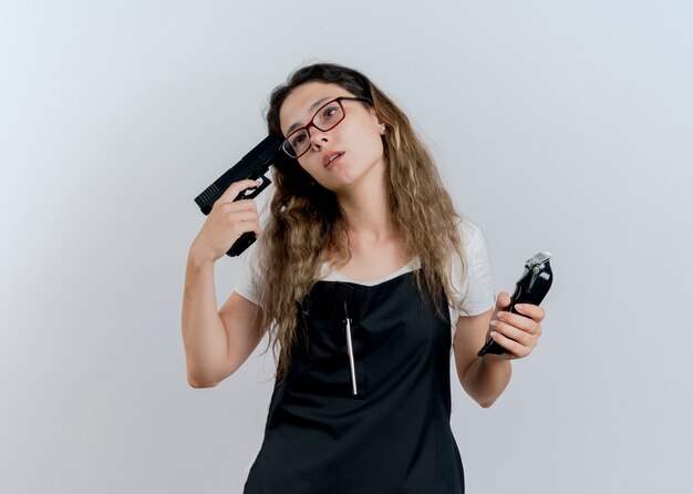 Young professional hairdresser woman in apron holding trimmer and pistol near her temple looking aside tired and bored standing over white wall