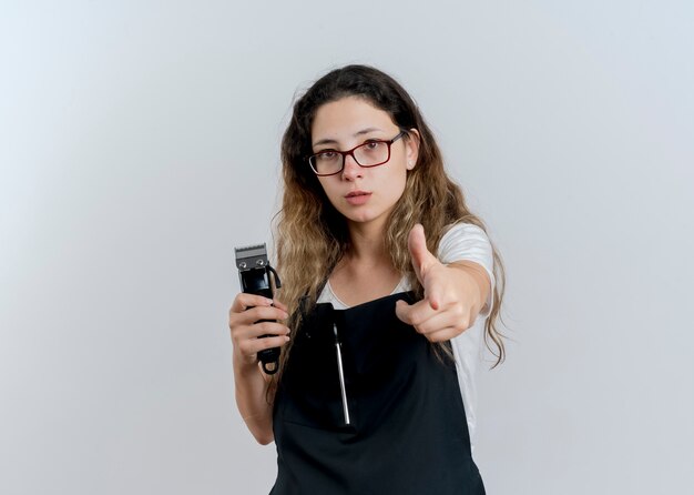 Young professional hairdresser woman in apron holding trimmer pointing with index finger at front looking confident standing over white wall