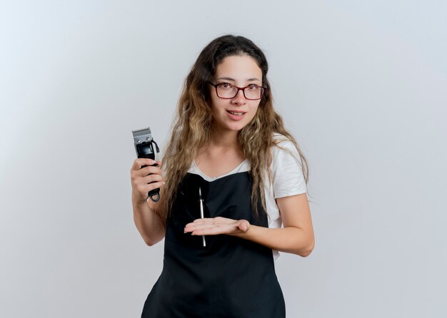 Young professional hairdresser woman in apron holding trimmer presenting it with arm of her hand smiling standing over white wall