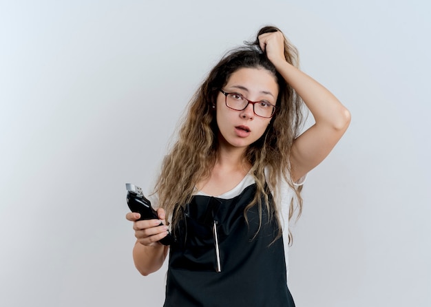 Young professional hairdresser woman in apron holding trimmer touching her hair looking confused standing over white wall