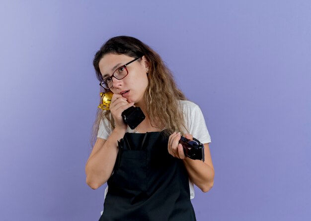 Young professional hairdresser woman in apron holding trimmer and trophy looking aside puzzled 