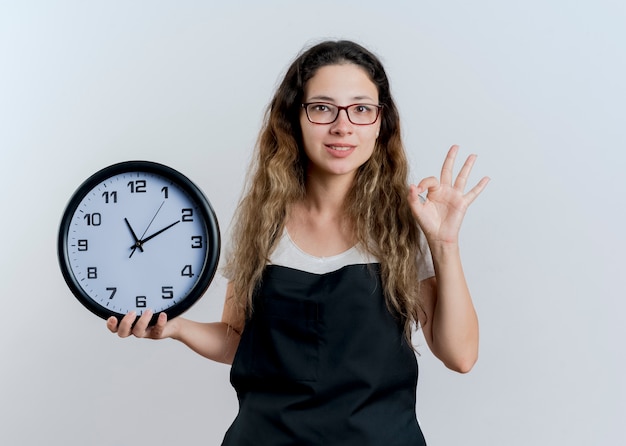 白い壁の上に立っているOKのサインを示す正面を見て壁時計を保持しているエプロンの若いプロの美容師の女性