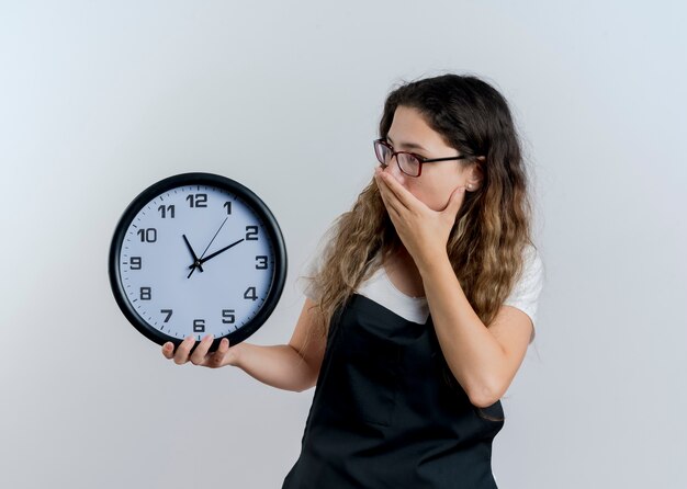 Young professional hairdresser woman in apron holding wall clock looking at it covering mouth with hand being shocked standing over white wall