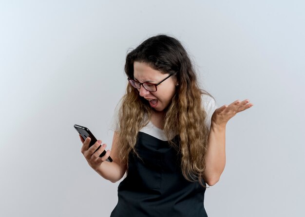 Young professional hairdresser woman in apron looking at her smartphone screen going wild shouting with aggressive expression standing over white wall