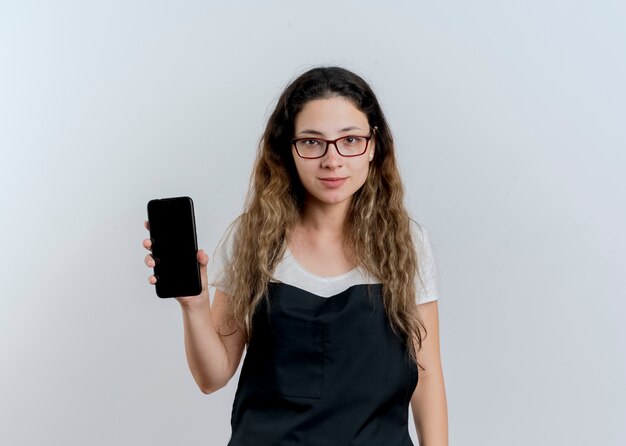 Молодой профессиональный парикмахер женщина в фартуке показывает смартфон, глядя на переднюю улыбку уверенно стоя над белой стеной