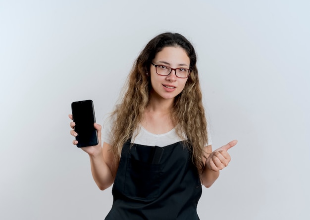Молодой профессиональный парикмахер женщина в фартуке показывает смартфон улыбается, глядя на перед, показывает палец вверх, стоя над белой стеной