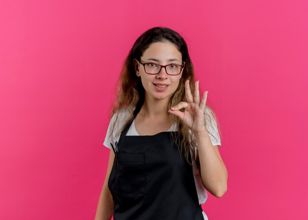 Young professional hairdresser woman in apron smiling doing ok sign 